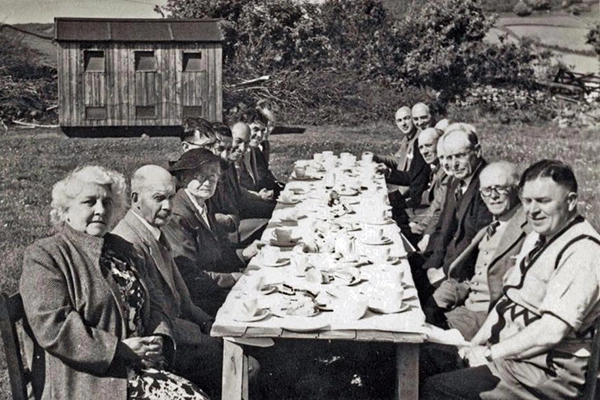 Black and white historic photograph of a community gathering at The Community Hall Grizebeck, with people seated at a long table outdoors