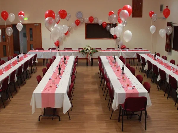 Interior of Grizebeck Community Hall prepared for a celebration, with long tables laid with tablecloths, dishes and balloons before guests arrive