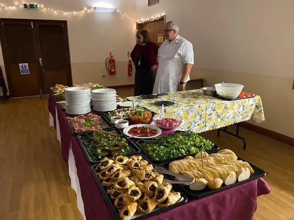 Two people standing beside tables of food prepared for a Greek food night at the Community Hall Grizebeck